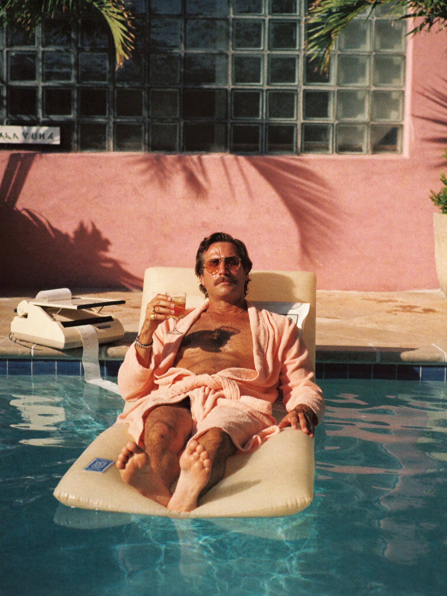 A man in a pink bathrobe lies on a pool float with a drink, beside a fax machine printing into the pool.